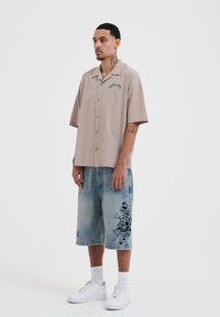 Korte mouwen beige overhemd met borduurwerk, gecombineerd met lichtblauwe denim Bermuda-shorts met zwarte grafische ontwerpen en sneakers.