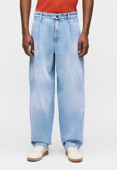 Άνδρας που φοράει ανοιχτό μπλε jeans με φαρδιά μπατζάκια, λευκά αθλητικά παπούτσια και κόκκινο πουκάμισο, στέκεται με τα χέρια χαλαρά δίπλα στο σώμα.