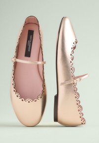 Chaussures plates Mary Jane en or métallique avec des bords perforés en forme d'ondulations et une fine bride à boucle sur fond vert clair.
