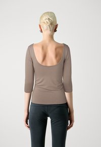 Brown fitted top with three-quarter sleeves and a wide scoop back, made from smooth material. Paired with dark, form-fitting trousers.
