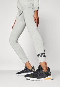 Leggings gris avec un logo "PUMA" noir à la cheville ; assortis avec des chaussures de sport noires avec des accents jaunes et une finition texturée.