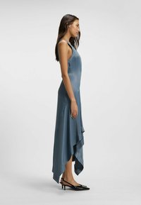 Satin, ärmelloses, asymmetrisches Kleid in Schieferblau mit fließendem Saum, kombiniert mit schwarzen spitzen Pumps.