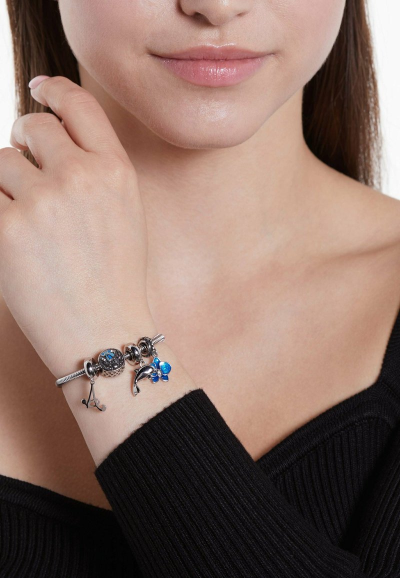 Zilveren armband met blauwe en zilveren bedels, gestructureerde kralen en een dolfijn. Draag gedragen om een pols met een zwarte geribbelde trui.