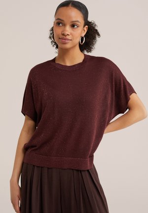 Femme portant un haut en maille pailletée bordeaux à manches courtes et une jupe plissée marron foncé, debout avec une main sur la hanche, regardant légèrement vers la droite.