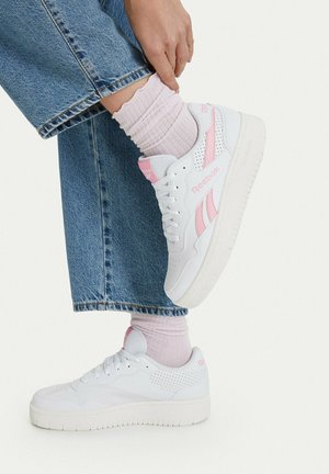 Person iført hvide og pink Reebok-sneakers, lyserøde sokker og blå denimbukser, løfter den ene fod for at røre anklen med hånden.