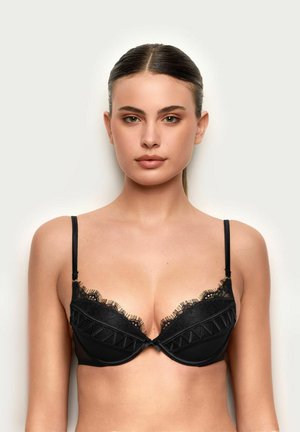 Frau mit glattem, dunklem Haar in einem schwarzen BH mit Spitzenbesatz, die frontal vor einem einfarbigen hellen Hintergrund steht.