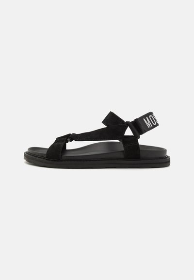 Zwarte sandaal van suede en rubber, met kruislings geïnterde banden en een brede verstelbare enkelband met het woord "MOSS" in het wit.