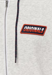 Felpa zip a collo alto grigio chiaro con un patch nero e rosso "ORIGINALS" sul petto, caratterizzata da un cappuccio con lacci neri intrecciati e dettagli rossi.