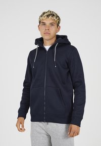 Marine blauwe zip-up hoodie van zacht materiaal, met een voorzak, een capuchon met trekkoord en geribbelde mouwen. Model draagt grijze joggingbroek.