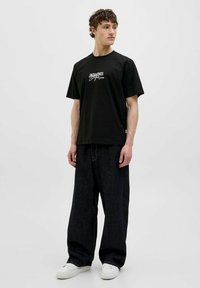 Sort bomulds T-shirt med hvidt logo, parret med løse mørke denimbukser og hvide sneakers. Modellen står afslappet op ad en lys baggrund.