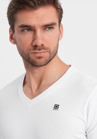 Weiße Baumwoll-V-Ausschnitt-T-Shirt mit tailliertem Design, das ein kleines schwarzes Logo-Accessoire in der oberen linken Ecke aufweist. Weicher Textur.