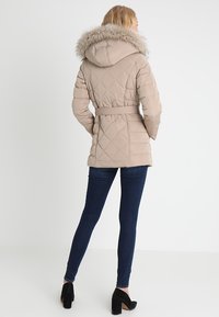 Béžová prošívaná péřová bunda s kapucí z umělé kožešiny, nastavitelným pásem v pase a diamantovým vzorem. Kombinována s tmavými džínami a černými lodičkami.