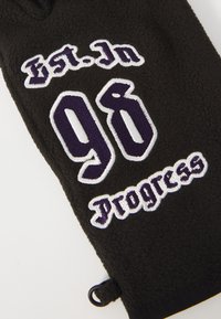 Tessuto in pile nero con testo ricamato in bianco e viola che dice "Est. In 96" e "Progress" con un piccolo anello nero sul bordo.
