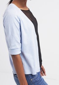 Ljusblå kardigan med uppvikt ärm, v-ringad design och mjuk textur, lager-på-lager över en svart mesh-top, i kombination med blå jeans.