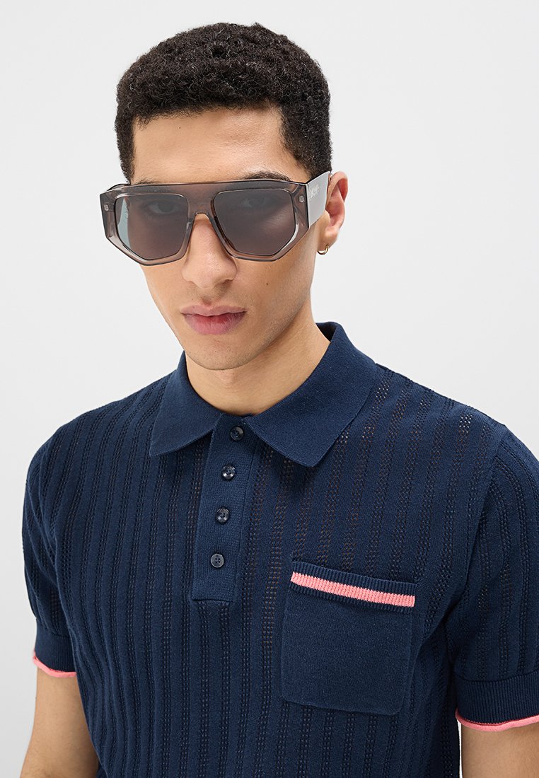 Polo en tricot bleu marine avec un motif texturé, trois boutons et une poche poitrine avec un bord rose ; le modèle porte des lunettes de soleil noires surdimensionnées.