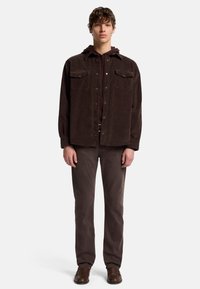 Giacca camicia in corduroy marrone con due tasche sul petto, indossata sopra una felpa bordeaux. Abbinata a pantaloni marroni scuri e scarpe marroni.