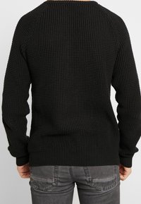 Homme portant un pull en tricot texturé noir et un jean gris foncé usé, vu de dos sur un fond uni.