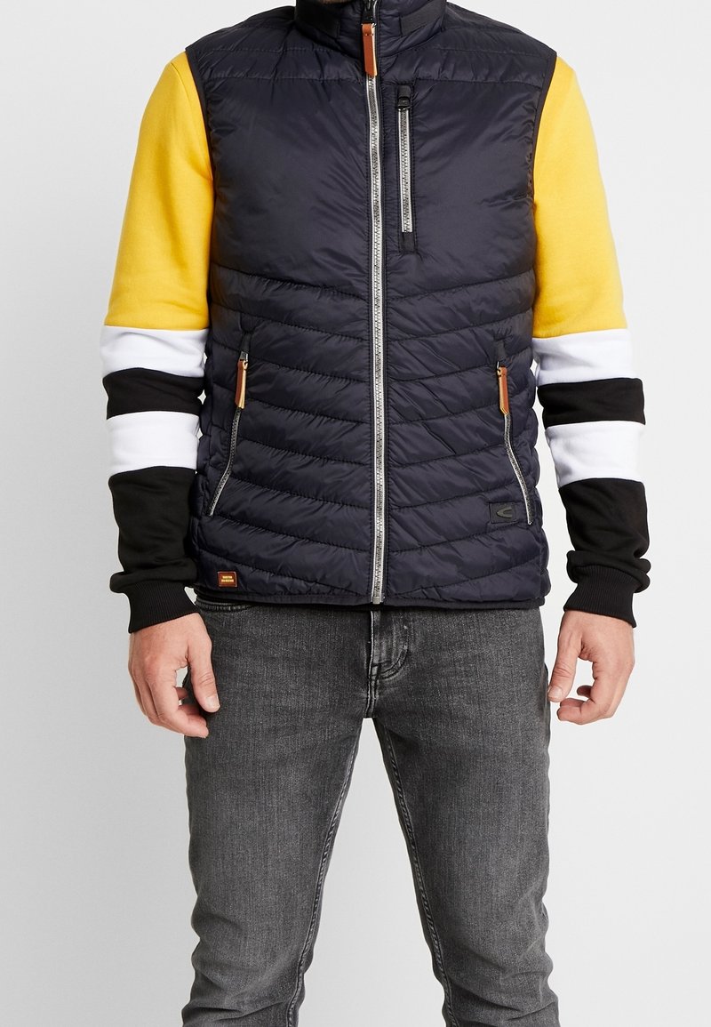 Homme portant un gilet matelassé noir avec des poches zippées par-dessus une chemise à manches longues avec des blocs de couleur jaune, blanc et noir et un jean gris.