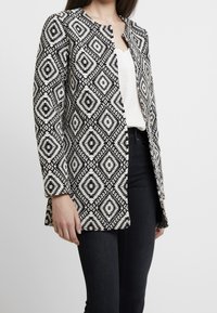 Giacca a righe in bianco e nero con un motivo a rombi, maniche lunghe, colletto arrotondato e apertura frontale, abbinata a una maglietta semplice e jeans scuri.