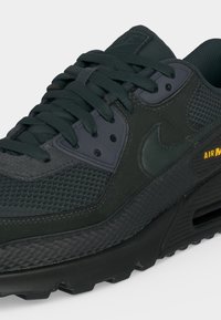 Μαύρο sneaker Nike Air Max με πάνελ από δίχτυ και δέρμα, κλείσιμο με κορδόνια, ορατή μονάδα αερόσολας και κίτρινο λογότυπο "Air Max" στο πλάι.