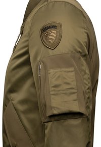 Giubbotto bomber verde oliva, realizzato in nylon morbido, presenta un ampio patch ricamato sulla spalla sinistra e una tasca con zip sul maniche.