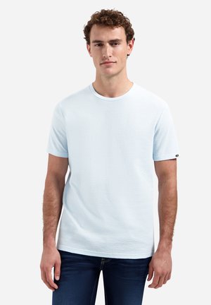 Man met krullend haar, gekleed in een lichtblauw, kortärmelig textuur T-shirt en donkerblauwe jeans, staand tegen een effen achtergrond.
