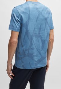 Blå t-shirt med korta ärmar som har en texturerad tyg med stora lövmönster i en mörkare blå nyans, rund halsringning och en avslappnad passform.