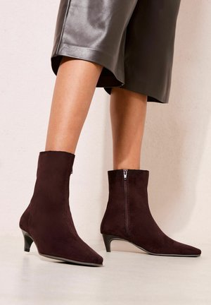 Kobieta nosząca ciemnobrązowe zamszowe botki za kostkę z ostro zakończonym noskiem i niskim obcasem, zestawione z krótką skórzaną spódnicą.