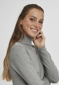 Pelēka turtleneck džemperis ar pieguļošu dizainu, maigu tekstūru un ribotām piedurknēm. Modelis pozē ar roku uz zoda pret baltu fonu.