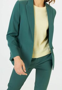 Veste turquoise et pantalon assorti, présentant des épaules structurées, un seul bouton, et un pull léger crème en dessous. Texture de tissu lisse.