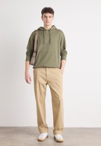 Felpa verde oliva con logo in rilievo, pantaloni beige e sneakers bianche. Il modello è in piedi su uno sfondo neutro, con le mani nelle tasche.