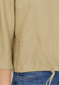 Beige lichtgewicht jack met een ronde zoom, korte mouwen en subtiele textuur; voorzien van een verstelbaar trekkoord in de taille.