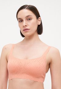 Bralette de cor pêssego feito de um material texturizado e elástico com um padrão geométrico e bordas recortadas. Alças largas e decote em forma de U.