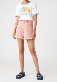 Persona vistiendo una camiseta blanca con un logo azul y amarillo, pantalones cortos de mezclilla rasgados rosas y sandalias de velcro azules de pie sobre un fondo blanco.
