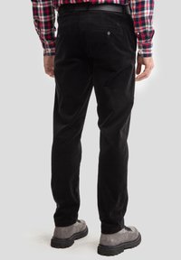 Pantalons en velours côtelé noir à coupe droite, avec deux poches arrière dotées de boutons en métal. Portés avec une chemise à carreaux et des bottes grises.