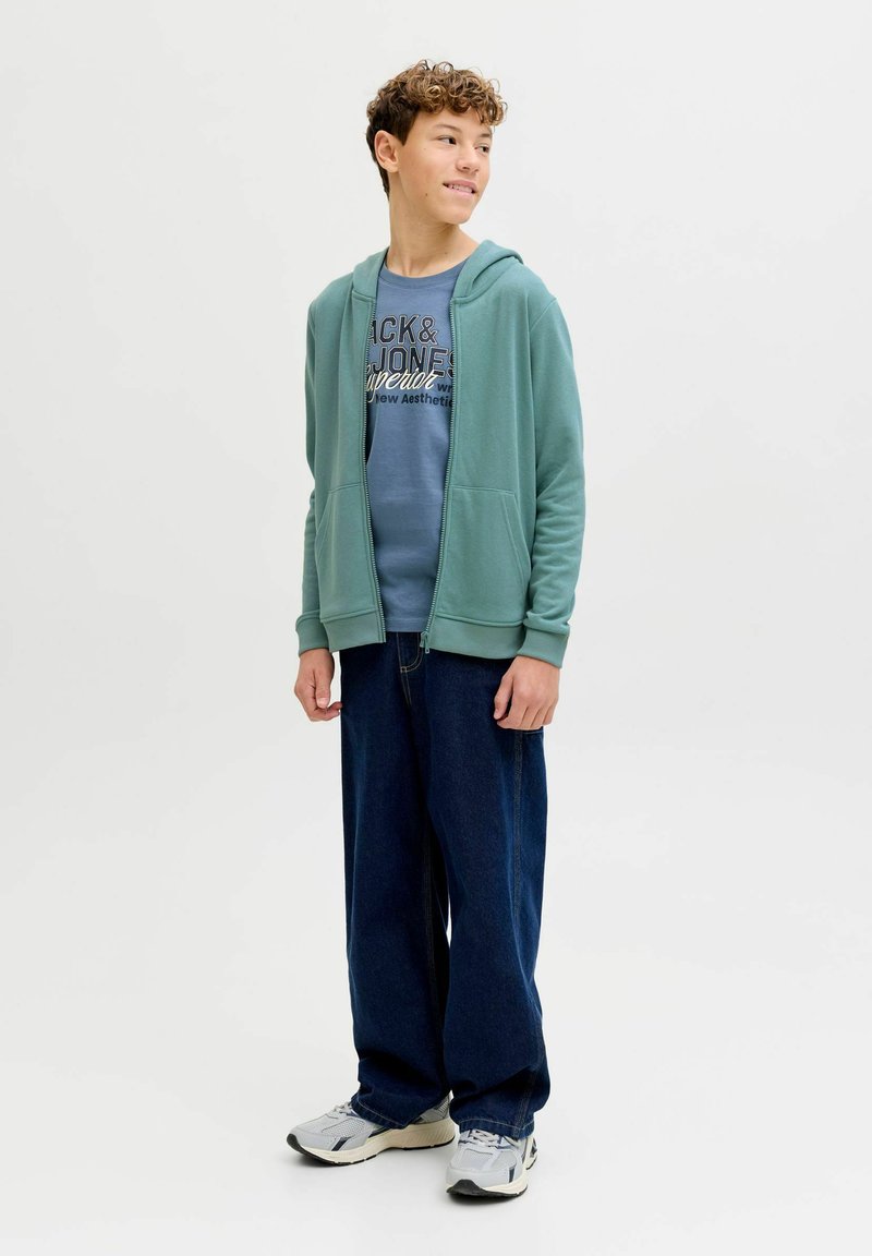 Lichtgroene hoodie met ritssluiting over een blauw grafisch T-shirt, donkerblauwe jeans met brede pijpen en grijze sneakers. Krullend haar en een neutrale achtergrond.