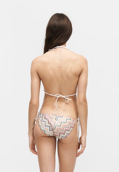 Γυναίκα εμφανίζεται από πίσω, φοράει πολύχρωμο κάτω μέρος μπικίνι με ζιγκ ζαγκ μοτίβο και λευκό ψηλόμεσο μπικίνι πάνω με δεμένα λουριά.