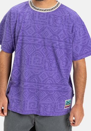 Homme portant un t-shirt violet ample avec des motifs géométriques abstraits et un petit patch rectangulaire montrant des palmiers sur le côté inférieur gauche.