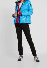 Doudoune bleu clair avec capuche, présentant un design matelassé, une fermeture éclair à l'avant et des accents noirs, superposée sur un top rose et un pantalon noir.