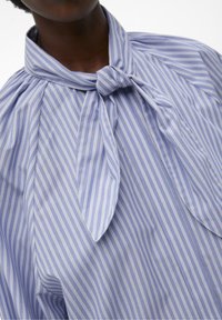 Camicia a righe azzurro chiaro e bianca con colletto annodato, caratterizzata da un tessuto liscio e dettagli arricciati vicino al collo.