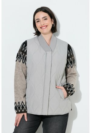 Smilende kvinde iført en lysegrå quiltet vest over en mønstret sweater med sorte og beige zigzag-designs, poserer med den ene hånd i lommen.