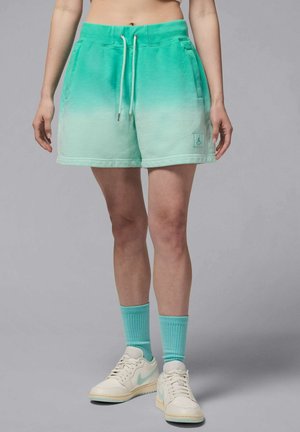 Henkilö, joka pukeutunut vihreisiin liukuvärjättyihin nyörishortseihin, vastaaviin puolipitkiin sukkiin ja valkoisiin lenkkareihin, joissa on vaaleanvihreitä yksityiskohtia, seisoo harmaata taustaa vasten.