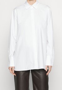 Camicia bianca di cotone con colletto classico, maniche lunghe, bottoni ai polsini e taglio dritto. Presenta un sottile dettaglio ricamato sul lato sinistro in basso.