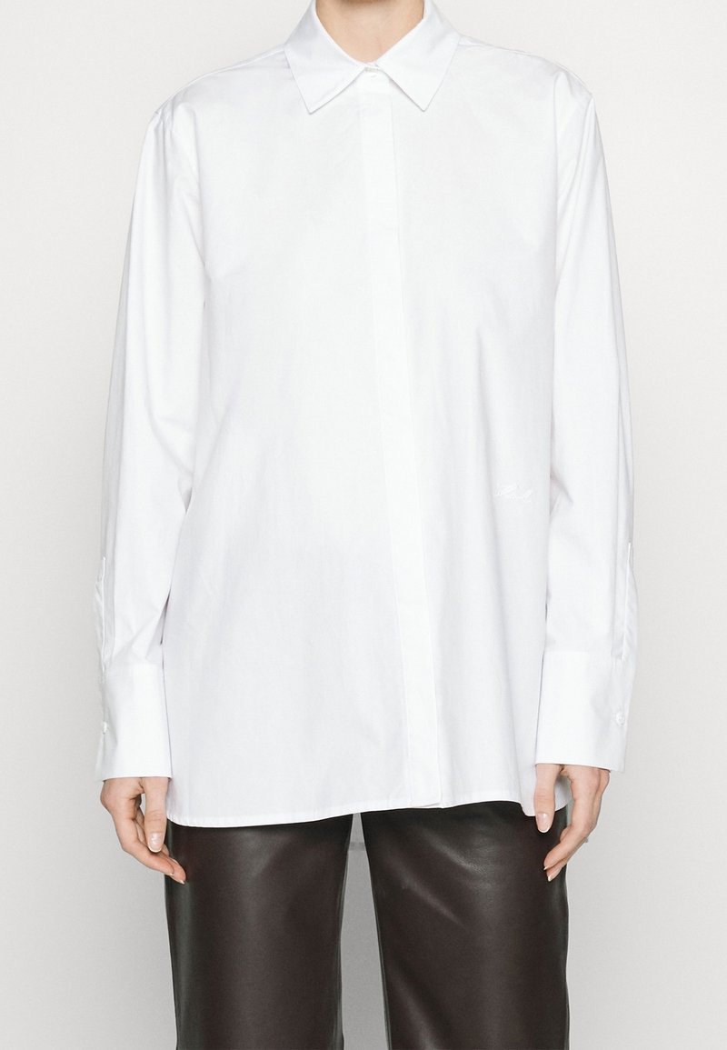 Camicia bianca di cotone con colletto classico, maniche lunghe, bottoni ai polsini e taglio dritto. Presenta un sottile dettaglio ricamato sul lato sinistro in basso.