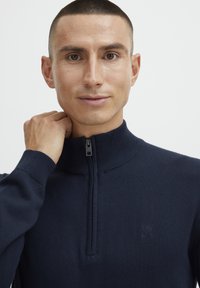 Sweater zippé marine avec un col montant, des poignets côtelés et un logo discret sur la poitrine. Texture lisse, design ajusté et matériau léger.