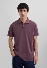 Camiseta polo de manga corta en algodón malva, con cuello clásico, tapeta de tres botones y un pequeño logotipo en el lado izquierdo del pecho.