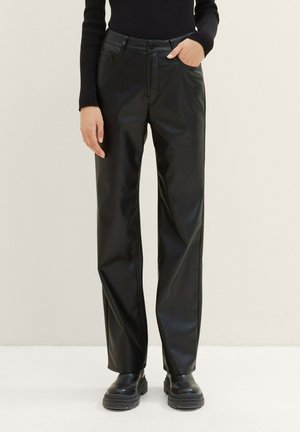 Persona que lleva pantalones rectos de cuero sintético negro, botas negras gruesas y una camiseta negra de manga larga, con una mano en el bolsillo.