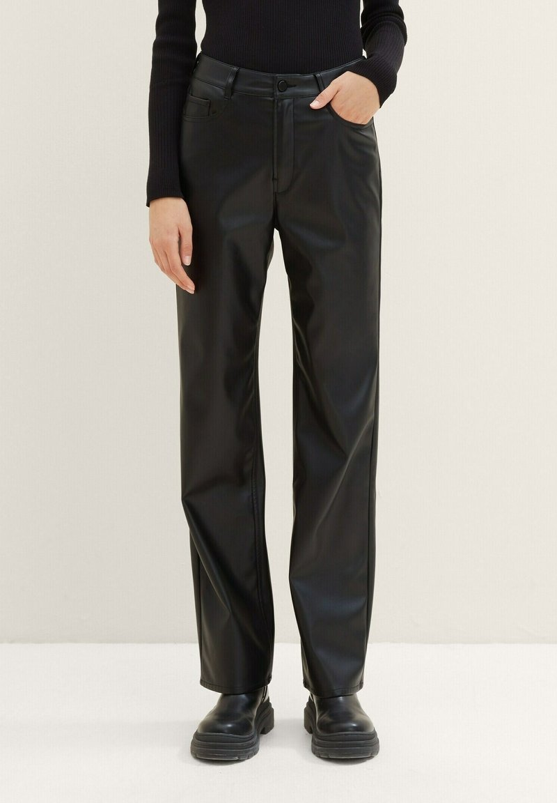 Personne portant un pantalon droit en cuir synthétique noir, des bottes épaisses noires et un haut noir à manches longues, une main dans la poche.