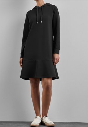 Femme portant une robe à capuche noire à manches longues et des baskets blanches, debout contre un mur gris uni.