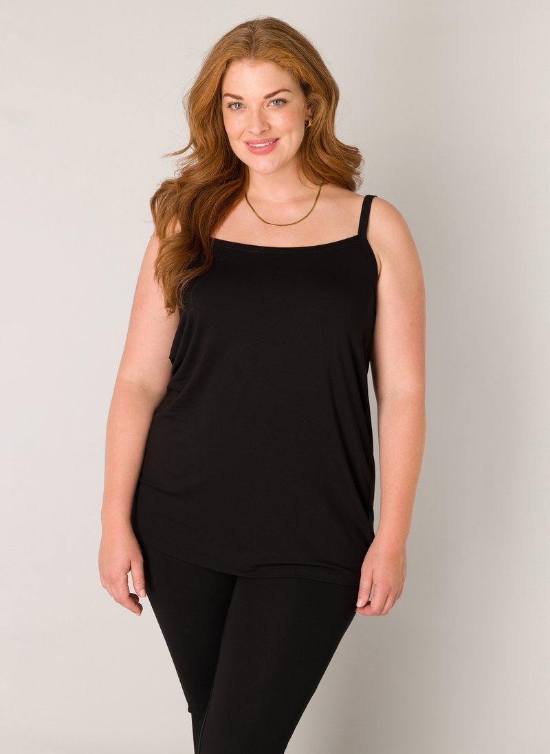 Lachende vrouw met lang rood haar, gekleed in een zwarte mouwloze top, zwarte legging en een gouden ketting, tegen een effen achtergrond.